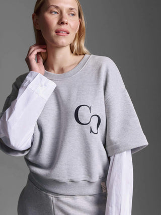 Junge Frau trägt das Cotton Candy Sweatshirt Manel grey melange, ein oversized, heather-graues Crewneck mit kurzen Ärmeln, über einem weißen Langarmhemd. Linke Brust zeigt ein schwarzes C- und S-Grafik.