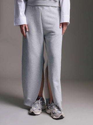 Die Cotton Candy Pant Mako grey/melan zeigt weite, hellgraue Sweatpants mit tiefen Schlitzen an den Beinen und off-white Sneakers, kombiniert mit einem dezenten weißen Shirt am Bund.