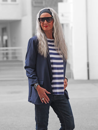 Erwachsene Person trägt den OFF#DLY Blazer Chiara marine über gestreiftem T-Shirt, kombiniert mit Jeans und Accessoires. Selbstbewusste Pose unterstreicht die lässige Eleganz des Blazers, perfekt für stilvolle Anlässe.
