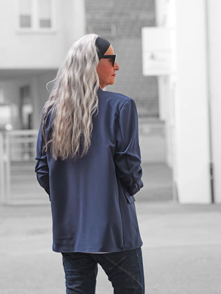 Person mit langem, silbrig-grauem Haar trägt den OFF#DLY Blazer Chiara marine, ein lässig-offenes Design in Dunkelblau, kombiniert mit legeren Jeans, während sie sich in einem urbanen Umfeld bewegt.