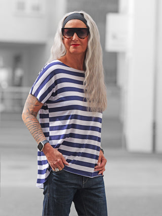 Eine Person trägt das OFF#DLY Marion Stripe Tee white/midblue, ein locker geschnittenes, gestreiftes Kurzarmshirt, kombiniert mit dunklen Jeans und Accessoires wie einer Smartwatch und einem Armband.