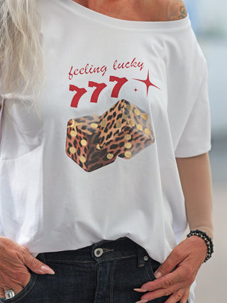 Person trägt OFF#DLY Tee LUCKY white mit rotem feeling lucky 777-Print und Leo-Würfeln. Lockerer Schnitt, off-shoulder Design, kombiniert mit dunkler Jeans und Schmuck.