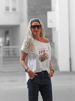 Frau trägt das OFF#DLY Tee LUCKY white, ein weißes, schulterfreies T-Shirt mit feeling lucky 777 und Leoparden-Würfeln. Kombiniert mit dunkler Jeans, Sonnenbrille und Armbanduhr.