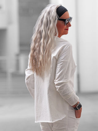 Person in weißem OFF#DLY Longsleeve Roma, lässiger Schnitt mit gerundetem Saum. Sichtbar: Brusttasche, Raw-Edge-Details. Langärmelig, natürliche Optik, ergänzt durch schwarze Sonnenbrille und Smartwatch im urbanen Hintergrund.