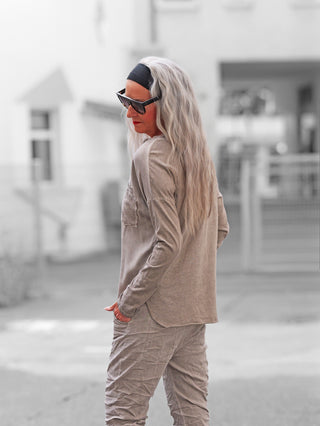 Person mit grauem, welligem Haar, trägt das OFF#DLY Longsleeve Roma taupe. Lockerer Schnitt, Brusttasche, leicht abgerundeter Saum. Hände in Taschen, dreht sich über die Schulter um.