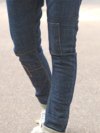 Nahaufnahme von Beinen in OFF#DLY Denim Cremona Dark Denim Jeans mit sichtbaren Patches und Kontrastnähten, kombiniert mit hellen Sneakers auf Asphalt, symbolisiert urbanen, lässigen Stil.