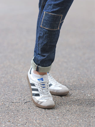 Unterer Teil von Beinen in hochgekrempelten OFF#DLY Denim Cremona Dark Denim Jeans, kombiniert mit Adidas Samba Sneakers, auf Bürgersteig stehend.