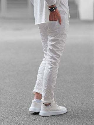 Person trägt OFF#DLY Joggpant Neapel offwhite, schmal geschnitten, leicht faltig. Hand in Gesäßtasche, kombiniert mit weißen Sneakers und einem quadratischen Smartwatch.
