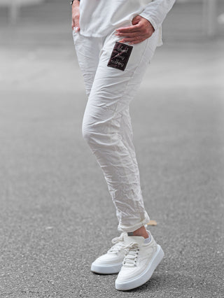 Nahaufnahme einer Person, die OFF#DLY Joggpant Neapel offwhite trägt. Die Hose zeigt einen dekorativen Patch „Sweet doing nothing“ auf dem Oberschenkel. Kombiniert mit weißen, klobigen Sneakers.