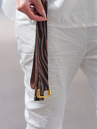 Die OFF#DLY Joggpant Neapel offwhite zeigt eine Hand mit einem schwarzen Zebra-Gürtel mit goldener Schnalle. Die weiße Hose betont Eleganz und Lässigkeit, perfekt kombiniert mit dem Gürtel.
