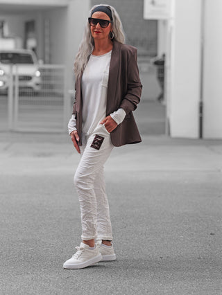 Person trägt OFF#DLY Joggpant Neapel offwhite, eine weiße, leicht geknitterte Hose mit Patch-Applikation auf dem Oberschenkel. Kombiniert mit Blazer, Sneakers und Accessoires für einen lässigen Look.