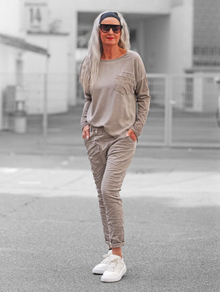 Person in OFF#DLY Joggpant Bari grey, taupe-beige, mit Crinkle-Optik, lässig-schmale Passform, kombiniert mit weißen Sneakers, steht entspannt auf gepflasterter Straße vor hellem Gebäude, Hände in Hosentaschen.