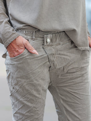 Nahaufnahme von OFF#DLY Joggpant Bari grey: Person trägt bequeme, leicht geknitterte Hose mit Five-Pocket-Stil und sichtbarem FASHION-Knopf, Hand in der Vordertasche. Ideal für entspannte, stilvolle Outfits.