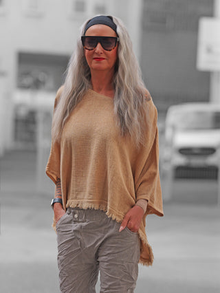 Erwachsene Person trägt das OFF#DLY Shirt Arianna camel, ein locker sitzendes, beige Fransen-Poncho, kombiniert mit hellgrauen, leicht geknitterten Hosen, während sie lässig auf einer Stadtstraße steht.