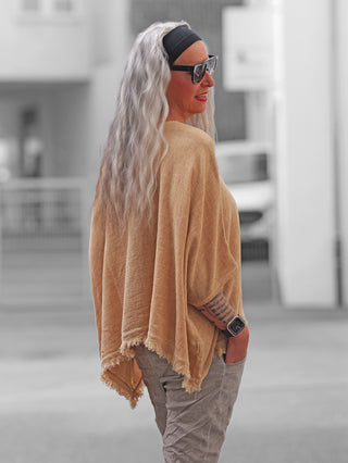 Die OFF#DLY Shirt Arianna camel zeigt eine Frau mit silbernem Haar in urbaner Umgebung, bekleidet mit einem locker sitzenden, fransenbesetzten Poncho, kombiniert mit grauen Hosen und schwarzem Haarband.