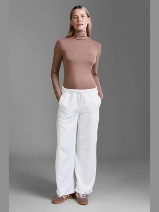 Person trägt Cotton Candy Pant Mehtab offwhite, eine locker geschnittene Cordhose mit Tunnelzug und geradem Bein, aus leichtem, geripptem Stoff, kombiniert mit einem braunen, eng anliegenden Rollkragenpullover.