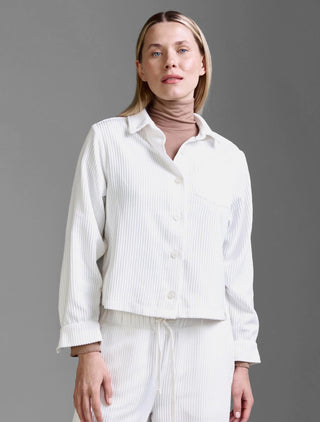 Eine Frau trägt das Cotton Candy Jacket Mesude offwhite, eine weiße, gerippte Cordjacke mit Knöpfen, über einem taubenfarbigen Rollkragenpullover, kombiniert mit passenden weißen, gerippten Cordhosen mit Kordelzug.
