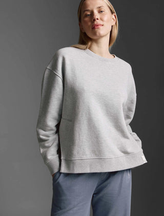 Person trägt das Cotton Candy Sweatshirt Manon grey melange, ein oversized, langärmeliges Rundhalssweatshirt mit offenen Seitennähten, kombiniert mit lässigen, slate-blauen Hosen. Hände in die Taschen gesteckt.