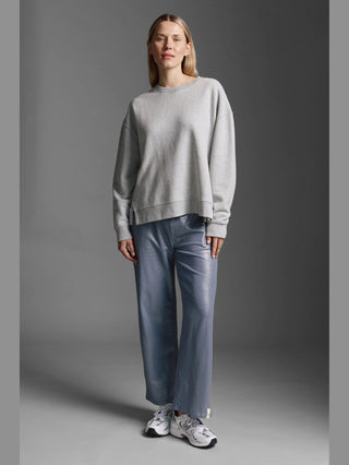 Frau trägt das Cotton Candy Sweatshirt Manon grey melange mit Rundhals und offenen Seitennähten. Es hat eine lässige, oversized Passform, kombiniert mit metallisch blauen Hosen und weißen Sneakers.