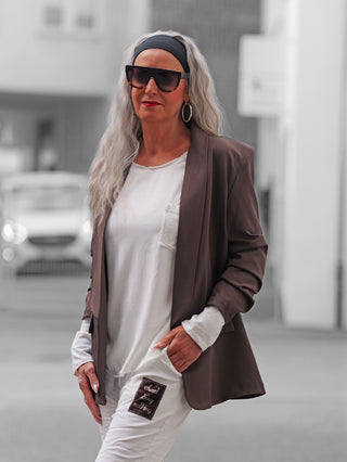 Ältere Frau im urbanen Umfeld trägt den OFF#DLY Blazer Eleonora Schoko, locker geschnitten mit gerafften 3/4-Ärmeln, über einem weißen T-Shirt und weißer Hose.
