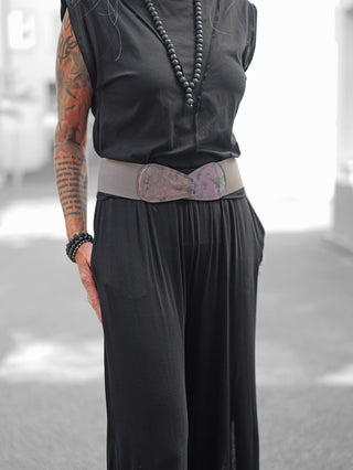 Person trägt OFF#DLY Gürtel Ornella anthrazit/metallic, elastischer Gürtel mit Leder-Schnallendetail, um eine lockere schwarze Kleidung. Armband und Halskette ergänzen den Look, Arm tätowiert.