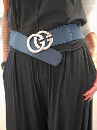 Nahaufnahme des OFF#DLY Gürtel GG denimblue/silber, getragen um die Taille eines schwarzen Kleidungsstücks, mit silberner GG-Schnalle und elastischem Design. Tattooierte Unterarme und Hände mit langen Nägeln sichtbar.