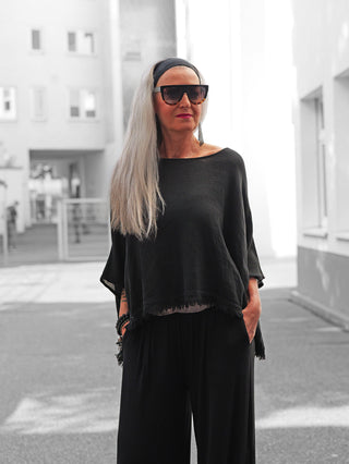 Mature Frau im Innenhof trägt OFF#DLY Shirt Arianna black, ein locker geschnittenes, schwarzes Oberteil mit fransigem Saum, kombiniert mit weiten Hosen. Sie hat silbernes Haar und trägt eine Sonnenbrille.