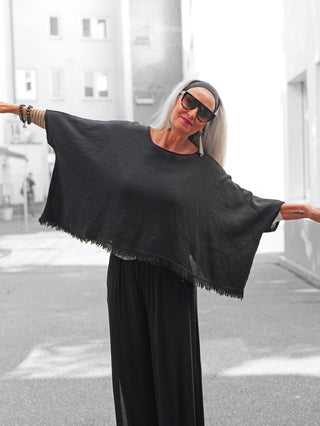 Person mit silbernem Haar trägt das OFF#DLY Shirt Arianna black, ein schwarzes, ponchoartiges Oberteil mit Fransen, in einer schmalen Stadtgasse. Kombiniert mit weiten, schwarzen Hosen.