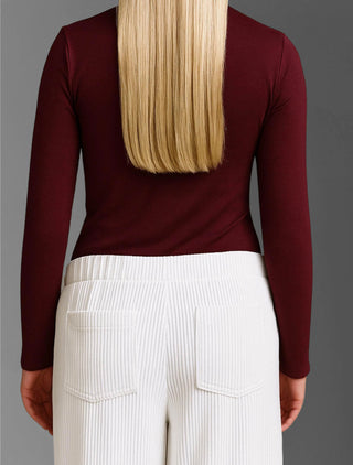 Rückansicht einer Person mit langen blonden Haaren, die das Cotton Candy Tee Blanche burgundy trägt, ein eng anliegendes Rollkragen-Langarmshirt, kombiniert mit hoch taillierten, weißen, gerippten Hosen.