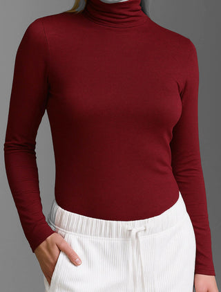 Person trägt das Cotton Candy Tee Blanche burgundy, ein eng anliegendes Langarmshirt mit Rollkragen, kombiniert mit weißen, gerippten Hosen. Ein Arm steckt lässig in der Hosentasche.