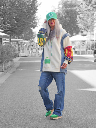 Person trägt OFF#DLY Sweatkleid Francesca creme mit auffälligen, bunten Grafik-Ärmeln, kontrastierenden Denim-Details und grünen Kordeln. Ideal für Retro-Liebhaber, in oversized Passform.
