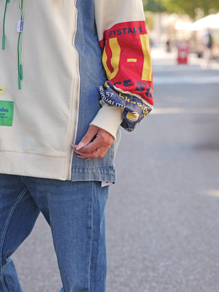 Person trägt OFF#DLY Sweatkleid Francesca creme mit grünen Kordeln, farbigen Print-Ärmeln und Denim-Details, kombiniert mit blauer Jeans. Nur unterer Torso und rechte Hand sichtbar.