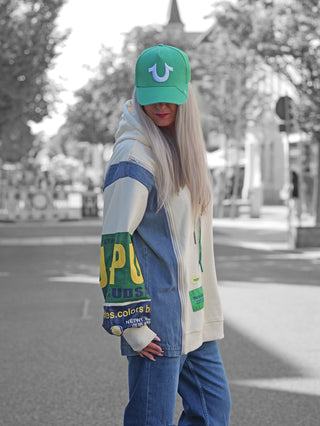 Junge Person trägt das OFF#DLY Sweatkleid Francesca creme mit auffälligen gummierten Ärmeldrucken, Denim-Einsätzen und Kapuze, kombiniert mit einer grünen Baseballkappe und blauer Jeans.
