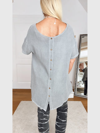 Person von hinten in lockerem, hellgrauem OFF#DLY Shirt Carlotta mit Holzknöpfen hinten, kombiniert mit dunklen Jeans mit Pinselstrichmuster. Kurze Ärmel, bequemer Schnitt.