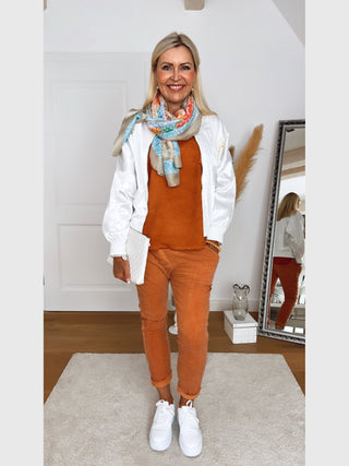 Frau trägt OFF#DLY Jacke Livia weiß mit schimmernder Oberfläche und dekorativen Patches. Kombiniert mit rostfarbenem Outfit, weißem Sneaker und farbigem Schal, hält sie eine weiße Clutch vor einem Spiegel.