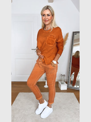 Frau trägt das OFF#DLY Longsleeve Roma rostorange, ein lässiges Oberteil mit Brusttasche und abgerundetem Saum, kombiniert mit passenden Hosen und weißen Sneakers vor einem Spiegel.
