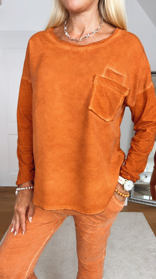 Person trägt OFF#DLY Longsleeve Roma rostorange, ein lässiges, langärmliges Oberteil mit Brusttasche und passender, schmal geschnittener Hose. Sichtbare Accessoires: Silberkette, Armbänder, weiße Uhr.
