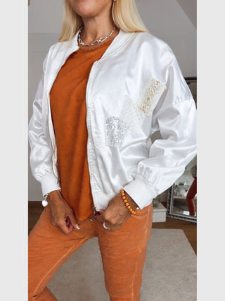 OFF#DLY Jacket Livia white, eine weiße, satinähnliche Bomberjacke mit dekorativen Spitzen- und Paillettenpatches, kombiniert mit einem lässigen, orangefarbenen T-Shirt und einer passenden Cordhose.