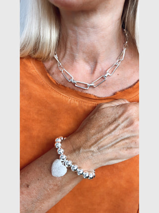 Nahaufnahme eines Halses mit einer OFF#DLY Gliederkette silber, einer massiven silbernen Kettenhalskette, getragen mit einem orangefarbenen Oberteil; dazu ein silbernes Armband mit Herzanhänger.