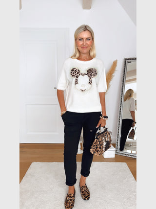 Frau in minimalistischem Raum trägt OFF#DLY Pulli Mickey creme mit großem Leo-Mickey-Motiv, kombiniert mit schwarzen Hosen, Leo-Loafers und passender Handtasche. Sie lächelt und trägt Schmuck.