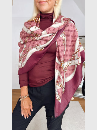 GUESS Noelle Scarf 130x130 WIN, ein großes quadratisches Tuch mit Kettengürtel-Print in Elfenbein und Gold, drapiert über burgunderfarbenem Rollkragenpullover, getragen mit goldfarbenen Kettenarmbändern.