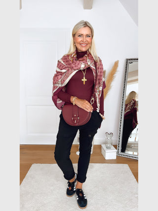 Mittelalterliche Frau in burgunderfarbenem Rollkragen, trägt GUESS Noelle Scarf 130x130 WIN mit Kettengürtel-Print, vor einem Spiegel stehend, schwarze Hose und Sneakers, goldene Accessoires.