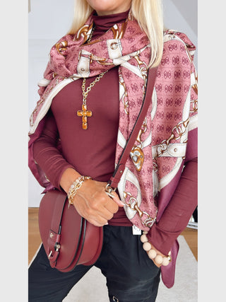 GUESS Noelle Scarf 130x130 WIN: Seidiger Schal in Rosa und Creme mit Ketten- und Schlossmotiven, elegant über burgunderfarbenem Oberteil drapiert, ergänzt durch goldene Accessoires und eine burgunderfarbene Umhängetasche.