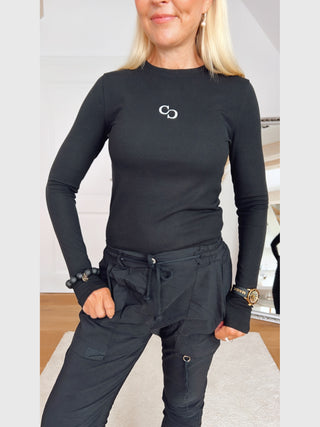 Person trägt das Cotton Candy Tee Lenja black: ein eng anliegendes, langärmeliges, geripptes Oberteil mit Rundhals und kleinem C-Logo auf der Brust. Kombiniert mit lässigen Cargo-Hosen.