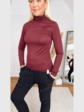 Frau trägt das Cotton Candy Tee Blanche burgundy, ein eng anliegendes, langärmeliges Rollkragenshirt, kombiniert mit schwarzen Cargo-Hosen. Sie hat Hände in den Taschen und trägt Perlenohrringe.