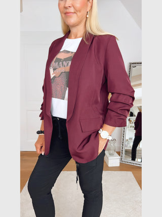 Person trägt den OFF#DLY Blazer Eleonora Bordeaux mit gerafften 3/4-Ärmeln über einem weißen Grafik-T-Shirt, kombiniert mit schwarzen Slim-Fit-Hosen. Accessoires: Uhr und Perlenarmbänder am linken Handgelenk.