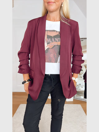 Person trägt den OFF#DLY Blazer Eleonora Bordeaux mit gerafften Ärmeln, über einem weißen T-Shirt. Der Blazer hat einen sportiv-eleganten Schnitt und fällt locker.