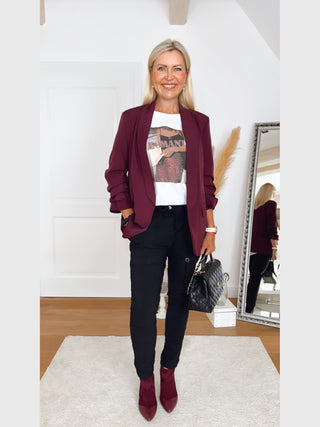 Frau in OFF#DLY Blazer Eleonora Bordeaux mit gerafften 3/4-Ärmeln, lockerem Schnitt, kombiniert mit schwarzer Hose und burgunderroten Stiefeletten, hält eine schwarze Tasche vor einem Spiegel.
