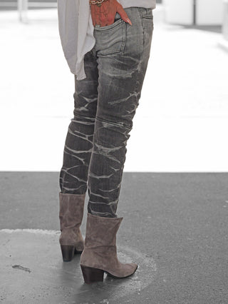 Person trägt OFF#DLY Denim Luino grey: graue, schmal geschnittene Jeans mit markanter Waschung, kombiniert mit taupefarbenen Stiefeln und einem goldfarbenen Armband. Hand ruht lässig in der Gesäßtasche.