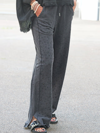 OFF#DLY Joggpant Daria black washed: Lose, schwarze Jogginghose mit Glitzernieten vorne, elastischem Bund und seitlichen Schlitzen am Beinabschluss, kombiniert mit schwarzen Sandalen und Perlenarmbändern.